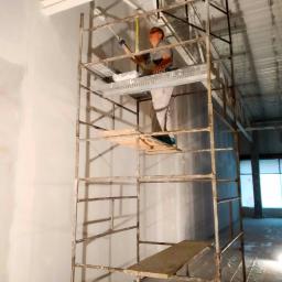 rewar.pl - Pracownik na rusztowaniu maluje ścianę w jasnym wnętrzu, widoczne niedoskonałości na powierzchni ściany przed malowaniem.