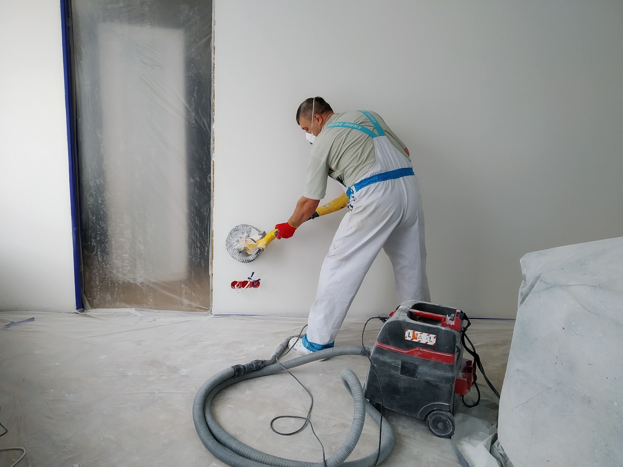 Pracownik w kombinezonie i masce szlifuje ścianę szlifierką z odkurzaczem, przygotowując ją do dalszych prac wykończeniowych; widoczne zabezpieczone folią drzwi.