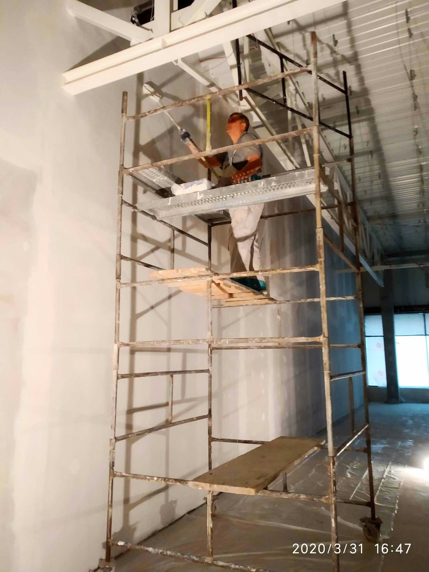 Pracownik na rusztowaniu maluje ścianę w jasnym wnętrzu, widoczne niedoskonałości na powierzchni ściany przed malowaniem.