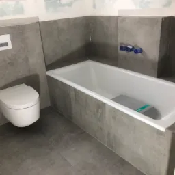 Wnętrze łazienki w trakcie prac wykończeniowych: szare płytki imitujące beton na ścianach i podłodze, biała wanna z ustawioną na niej stertą płytek, podwieszana toaleta z podtynkowym systemem...