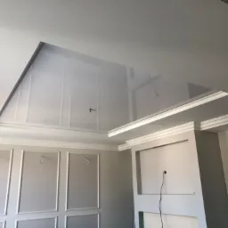 Wnętrze w trakcie wykańczania: ściany z dekoracyjnymi panelami i listwami, sufit podwieszany z wnęką, widoczne kable wystające ze ścian.