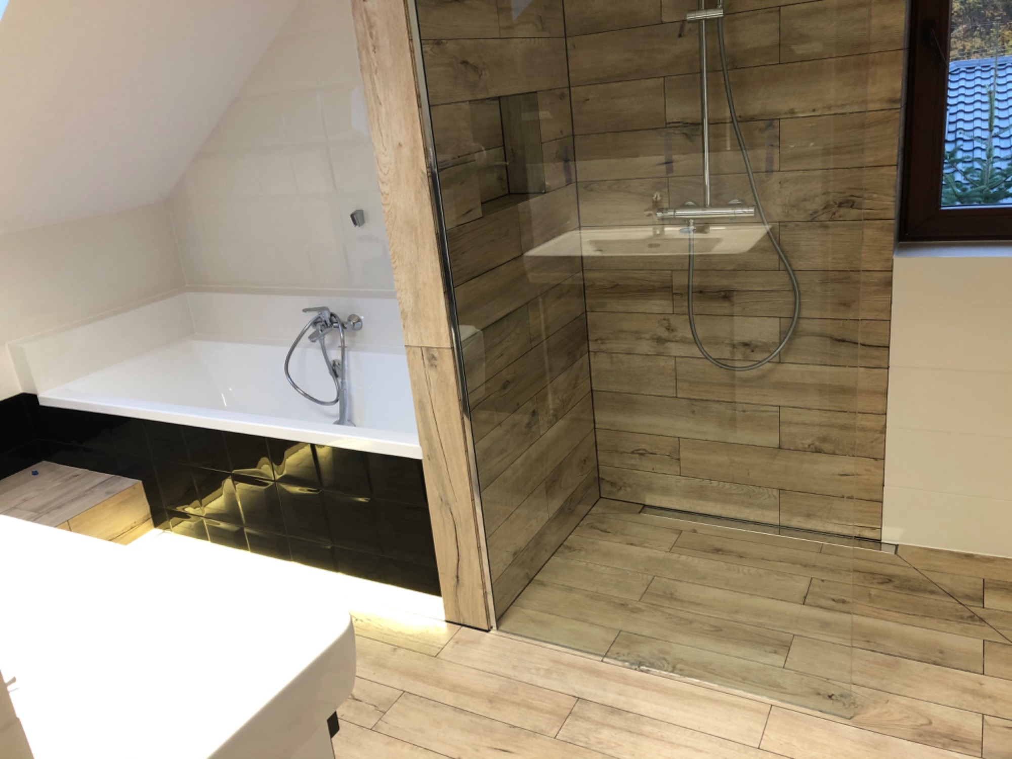 Nowoczesna łazienka na poddaszu z wanną obudowaną czarnymi płytkami 3D i kabiną prysznicową walk-in wyłożoną płytkami imitującymi drewno, widok na dachówkę przez okno.