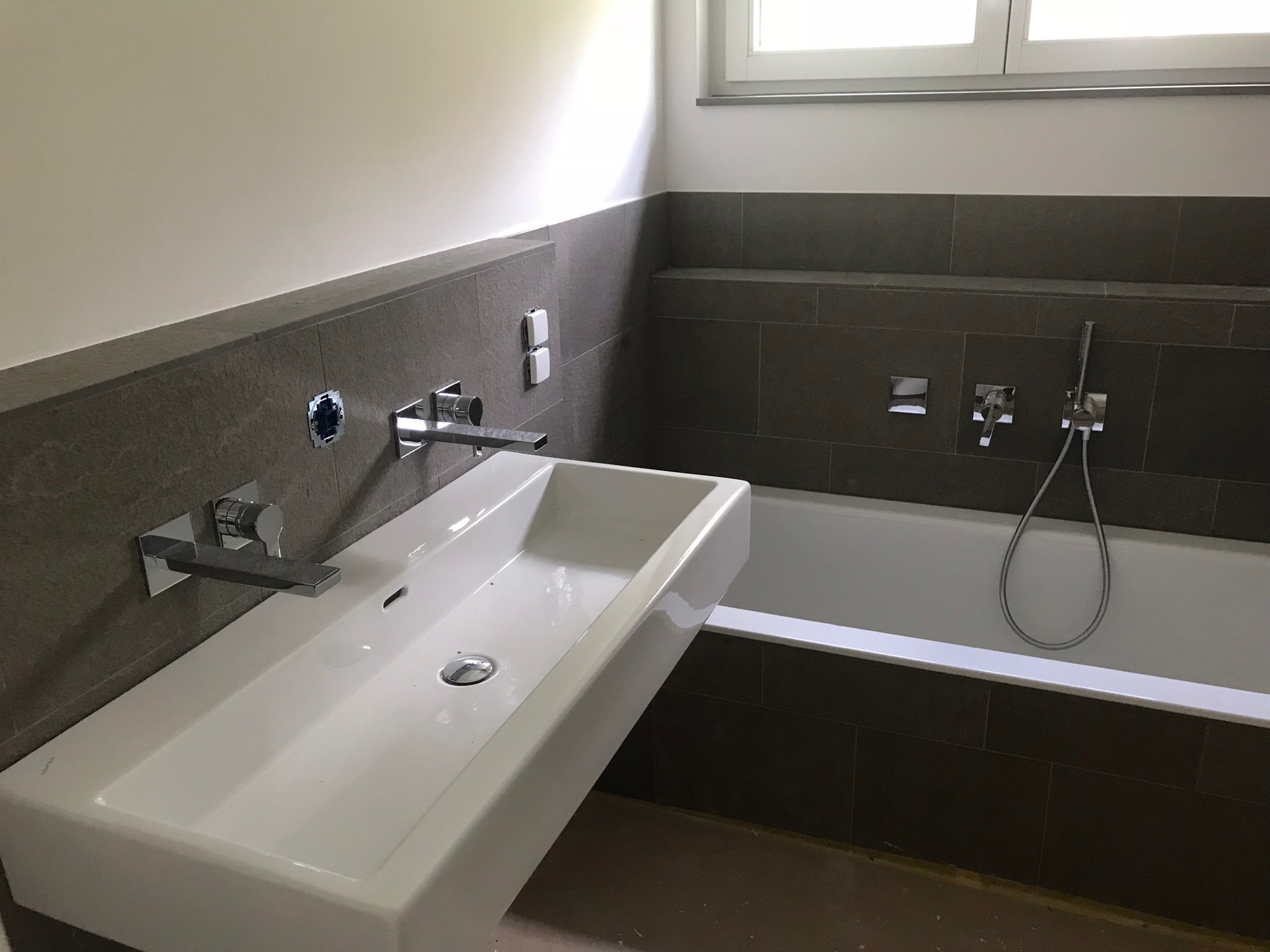 Nowoczesna łazienka z białą umywalką nablatową i baterią ścienną o kwadratowym kształcie, wykończona szarymi płytkami ceramicznymi, z widoczną wanną i oknem w tle.