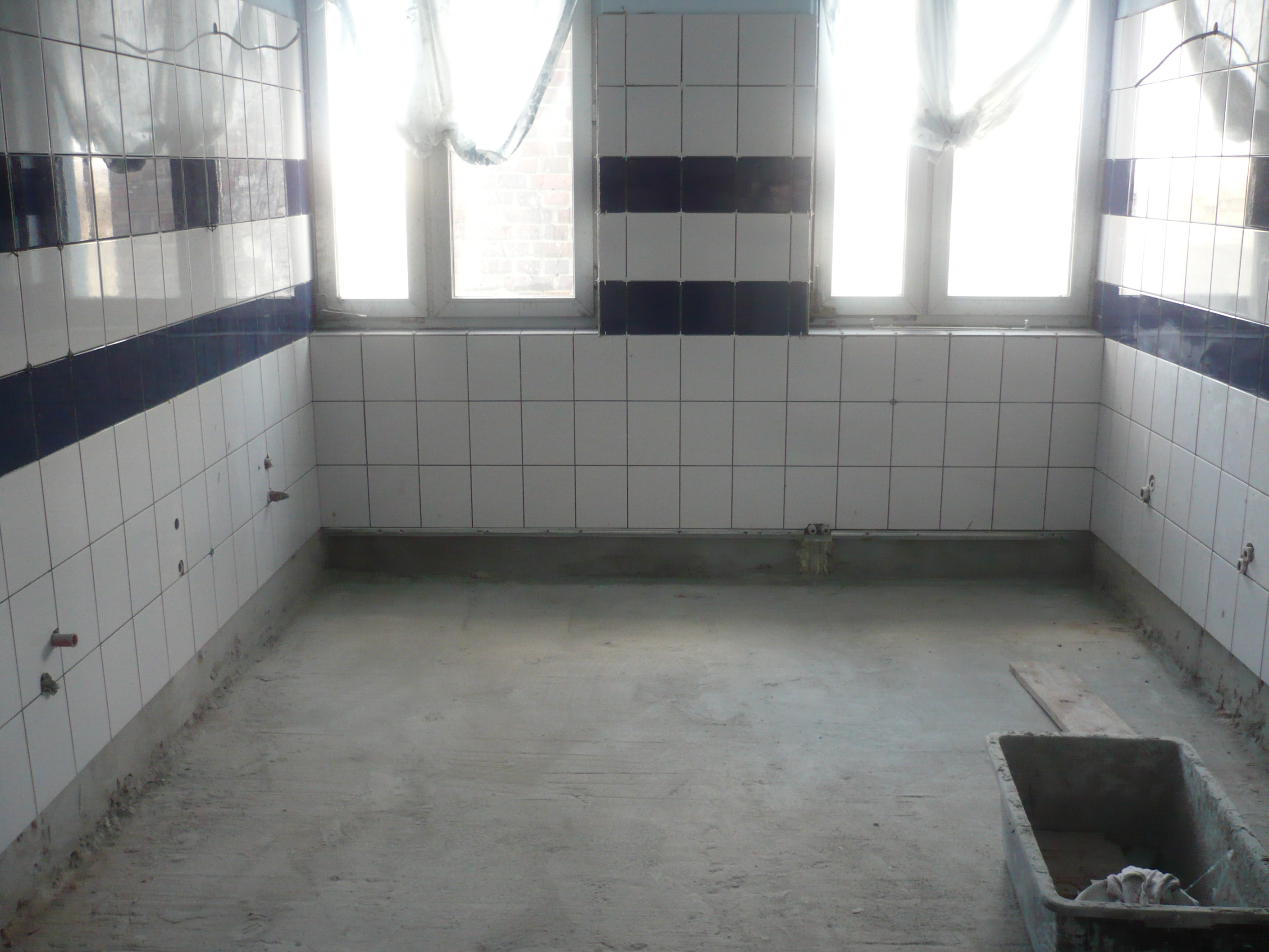 Wnętrze łazienki w trakcie remontu: białe płytki z ciemnoniebieskim pasem, surowy beton na podłodze, widoczne wystające rury instalacji wodnej, okno z zasłoniętą szybą i plastikowa kuweta na podłodze.