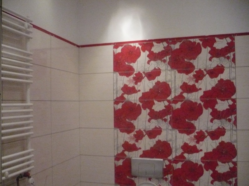 Wykończona łazienka z białymi płytkami, czerwonym akcentem kwiatowym na jednej ze ścian i grzejnikiem drabinkowym.