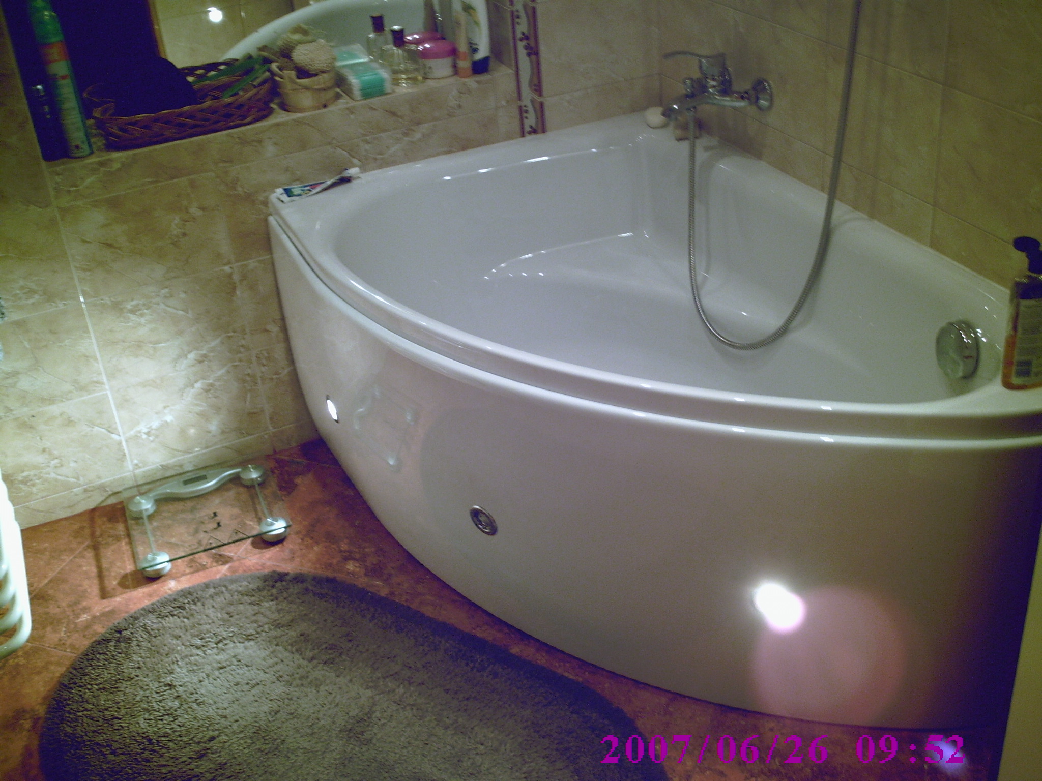 Narożna wanna z hydromasażem w łazience z beżowymi płytkami, widoczne dysze, bateria i słuchawka prysznicowa, obok waga łazienkowa i dywanik.