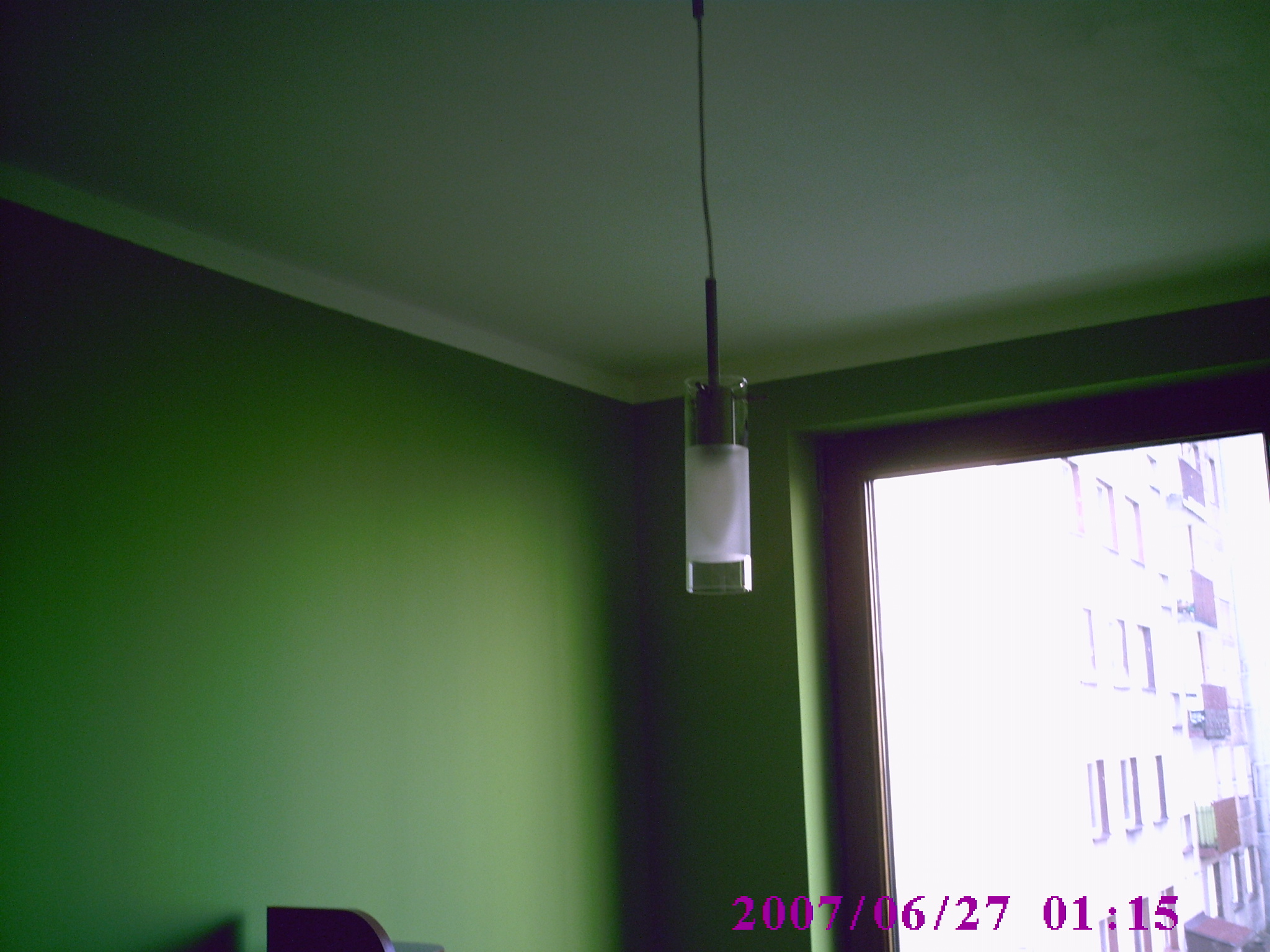 Lampa wisząca z cylindrycznym kloszem z mlecznego szkła, zawieszona na tle zielonej ściany i okna z widokiem na budynek mieszkalny.