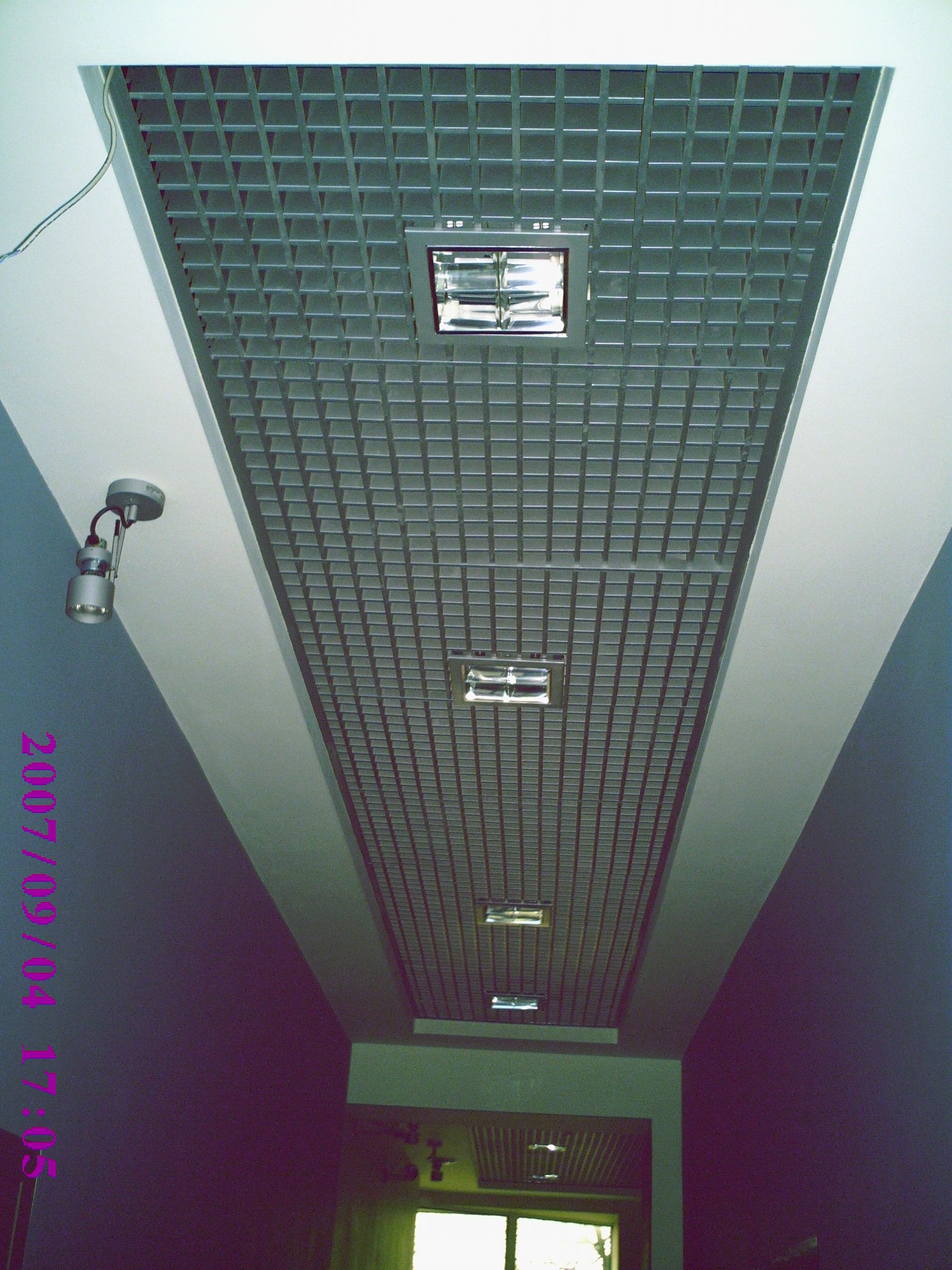 Sufit podwieszany z rastrem i linią lamp fluorescencyjnych w długim korytarzu, widok z dołu, z widoczną lampą punktową na ścianie i datą na zdjęciu.