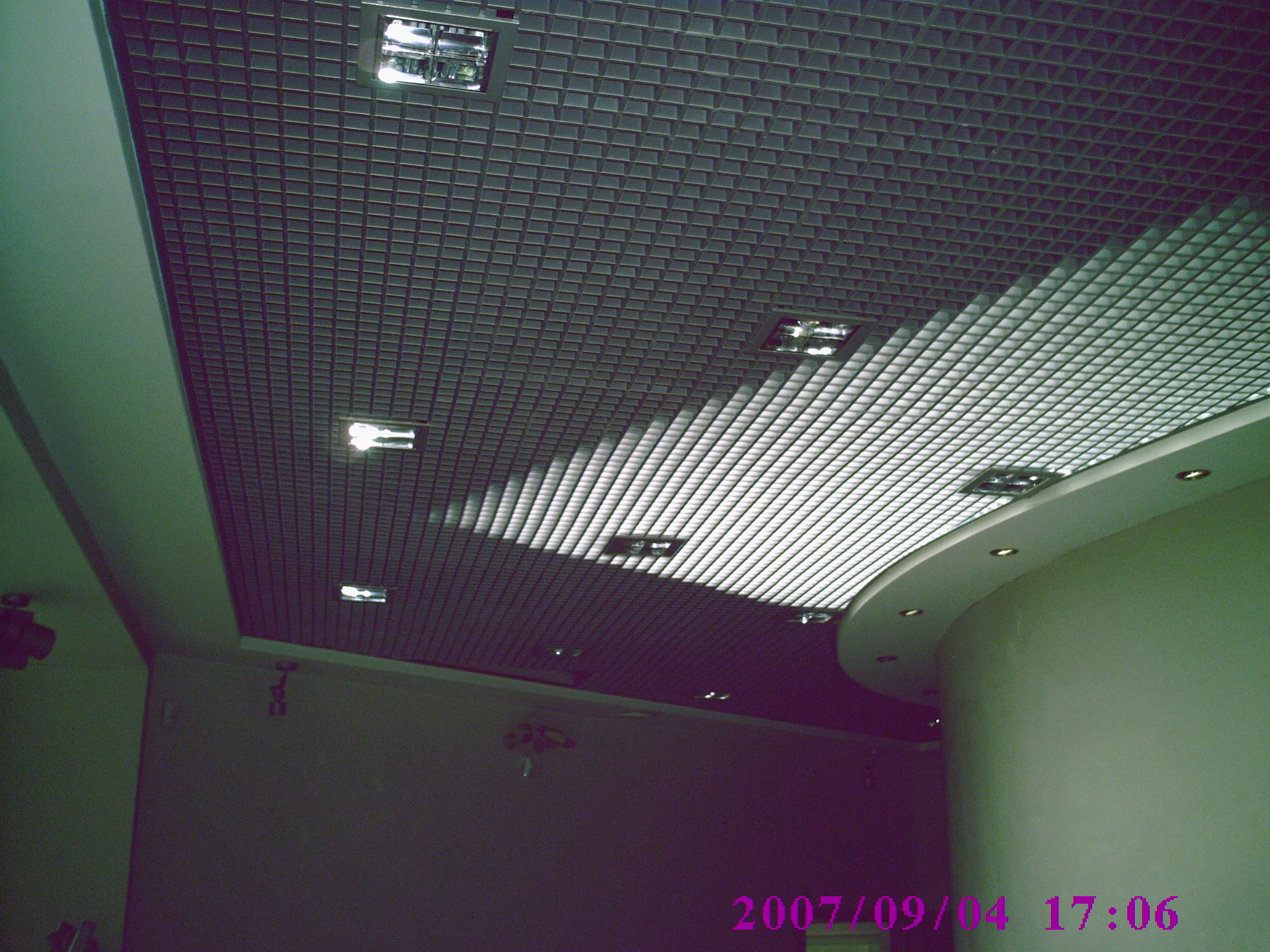 Sufit podwieszany z rastrem w kształcie kwadratów, oświetlony lampami wbudowanymi, z widocznym fragmentem zakrzywionego sufitu gipsowo-kartonowego z halogenami.