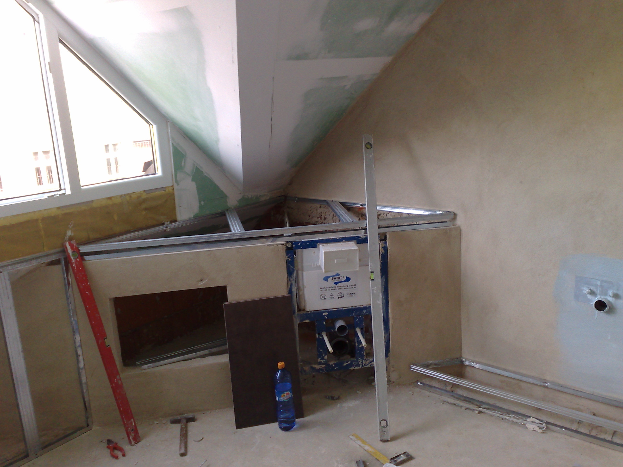 Montaż stelaża podtynkowego WC w łazience na poddaszu, widoczne profile metalowe, poziomica, płyty gipsowo-kartonowe i butelka wody.