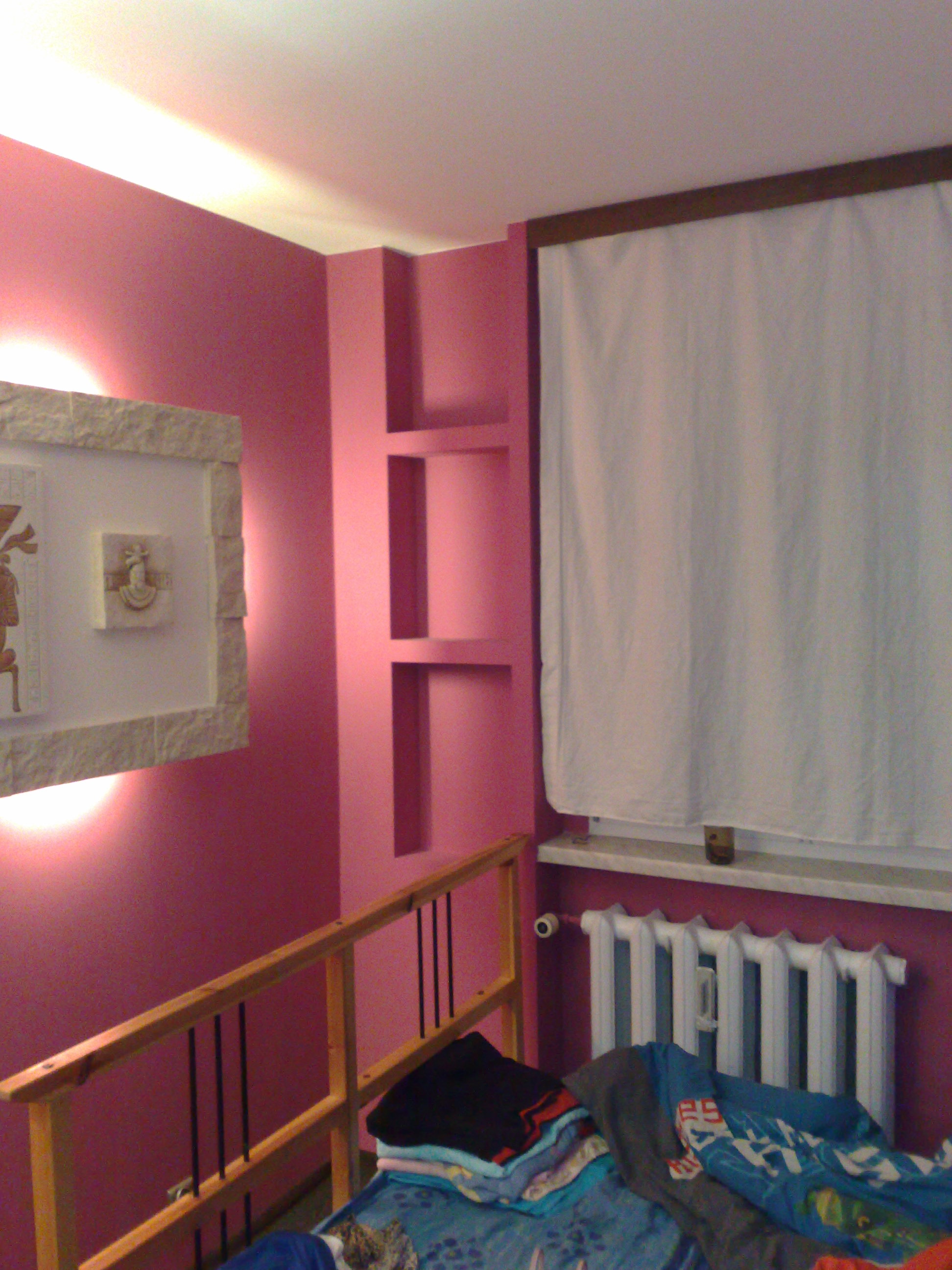 Różowa ściana w pokoju z wnękami, drewnianą barierką, białym grzejnikiem i zasłoną, z dekoracjami na ścianie podświetlonymi lampą.