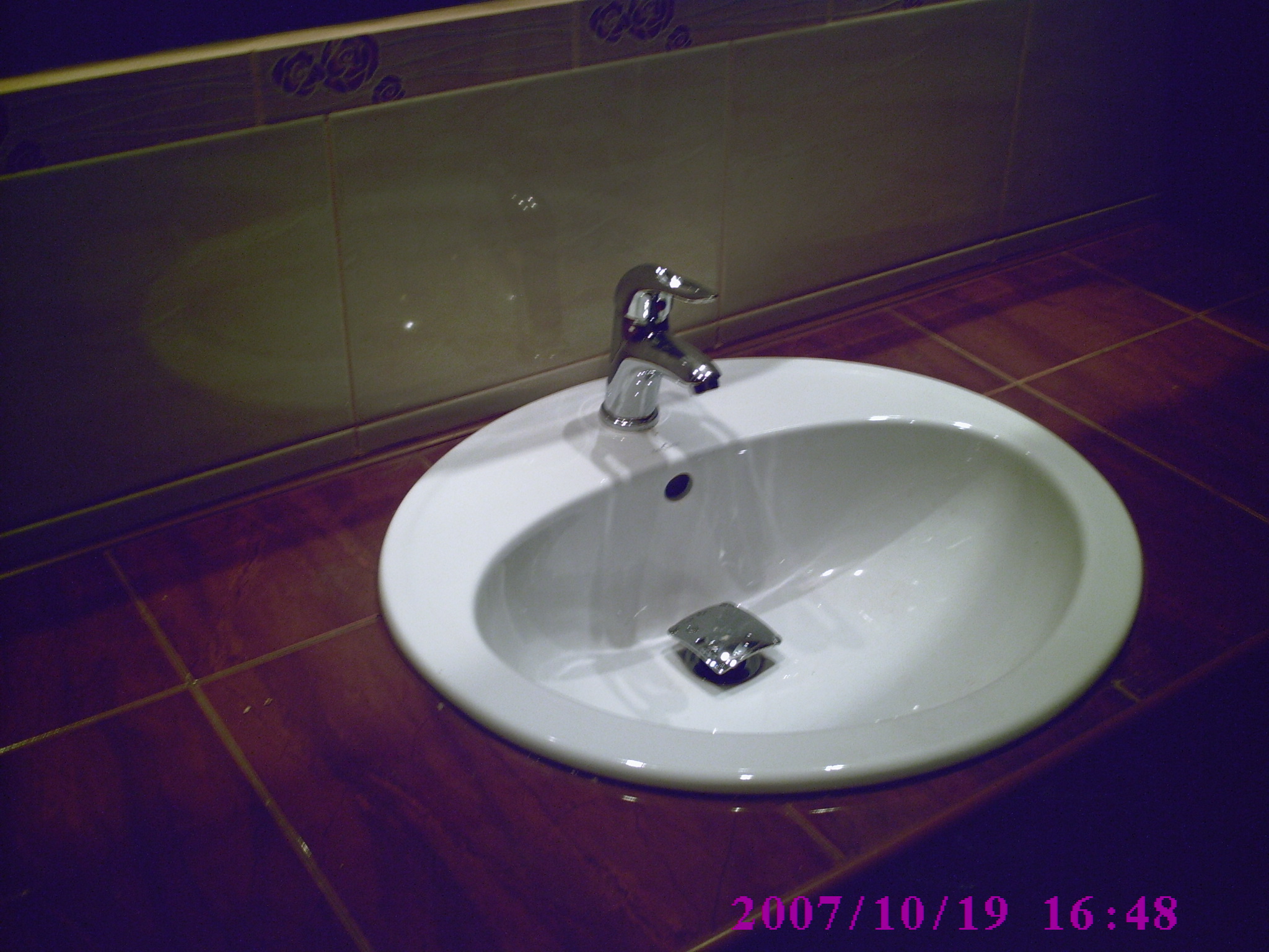 Biała umywalka z chromowaną baterią na brązowym blacie wyłożonym kafelkami. Na ścianie jasne płytki z delikatnym wzorem kwiatowym. Widoczny fragment daty i godziny zdjęcia.