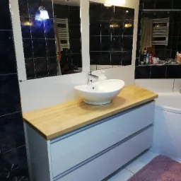 Nowoczesna łazienka z białą szafką pod umywalką z blatem z jasnego drewna, okrągła umywalka nablatowa, lustro w ramie i czarne płytki na ścianach.