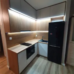 Nowoczesna kuchnia w odcieniach drewna i bieli z podświetleniem LED, czarną lodówką i drewnopodobną podłogą.