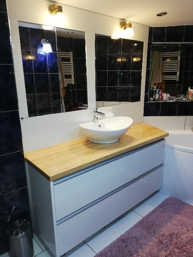 Nowoczesna łazienka z białą szafką pod umywalką z blatem z jasnego drewna, okrągła umywalka nablatowa, lustro w ramie i czarne płytki na ścianach.