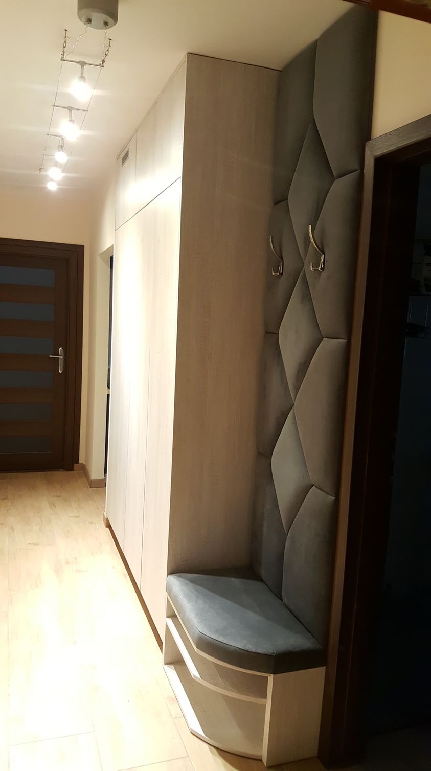 Wnętrze przedpokoju z zabudową: wysoka szafa z jasnego drewna, obok tapicerowany panel ścienny z wieszakami i siedziskiem z półkami na buty.