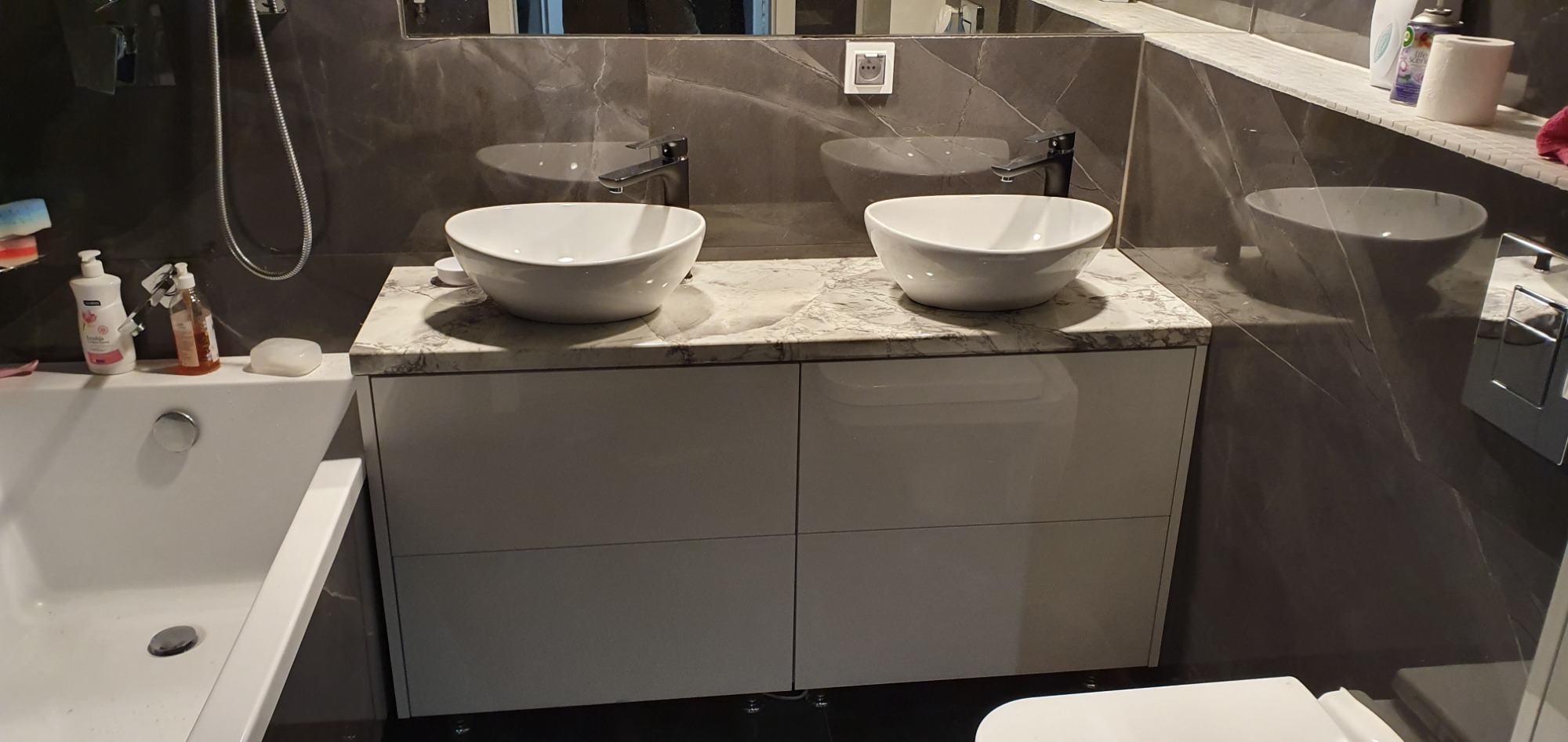 Nowoczesna łazienka z dwoma białymi umywalkami nablatowymi na szafce z marmurowym blatem, ciemne marmurowe ściany, minimalistyczna bateria.