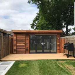 Nowoczesna, drewniana altana ogrodowa z dużymi, przeszklonymi drzwiami i tarasem z desek na tle zielonego trawnika.
