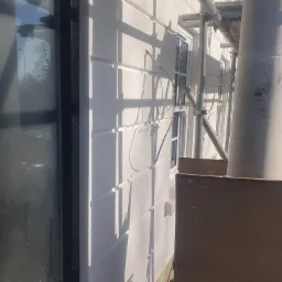 Fragment elewacji budynku w trakcie prac, widoczne rusztowanie, okno z ciemną ramą i świeżo otynkowana ściana z dekoracyjnymi listwami.