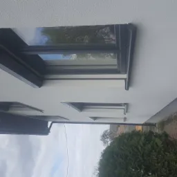 Fragment elewacji budynku z oknami o grafitowych ramach, widoczna faktura tynku baranka i odbicie drzewa w szybie okiennej.