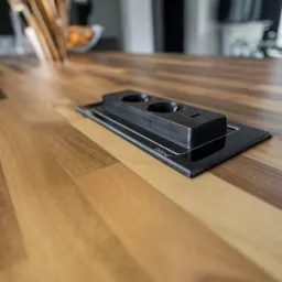 Gładki, drewniany blat z wbudowanym, czarnym gniazdem elektrycznym marki Evoline, widoczne dwa okrągłe gniazda i port USB, w tle rozmyte elementy wyposażenia kuchni.