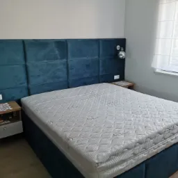 Sypialnia z łóżkiem tapicerowanym granatowym welurem, pikowany zagłówek z oświetleniem punktowym, biały materac i drewniane szafki nocne z ażurowymi serwetkami.