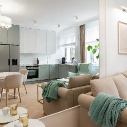 Jasna kuchnia z bladozielonymi szafkami i białymi blatami, widoczna z perspektywy salonu z beżową sofą i turkusowymi pledami, okrągły stół z krzesłami w tle.