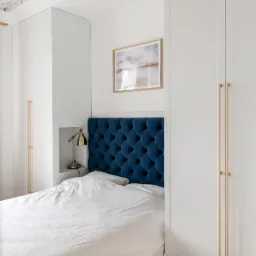 Biała szafa wnękowa z długimi, złotymi uchwytami, wkomponowana we wnękę nad łóżkiem z granatowym, pikowanym zagłówkiem i lampką nocną, na tle ściany z obrazem i sufitu z motywem kwiatowym.