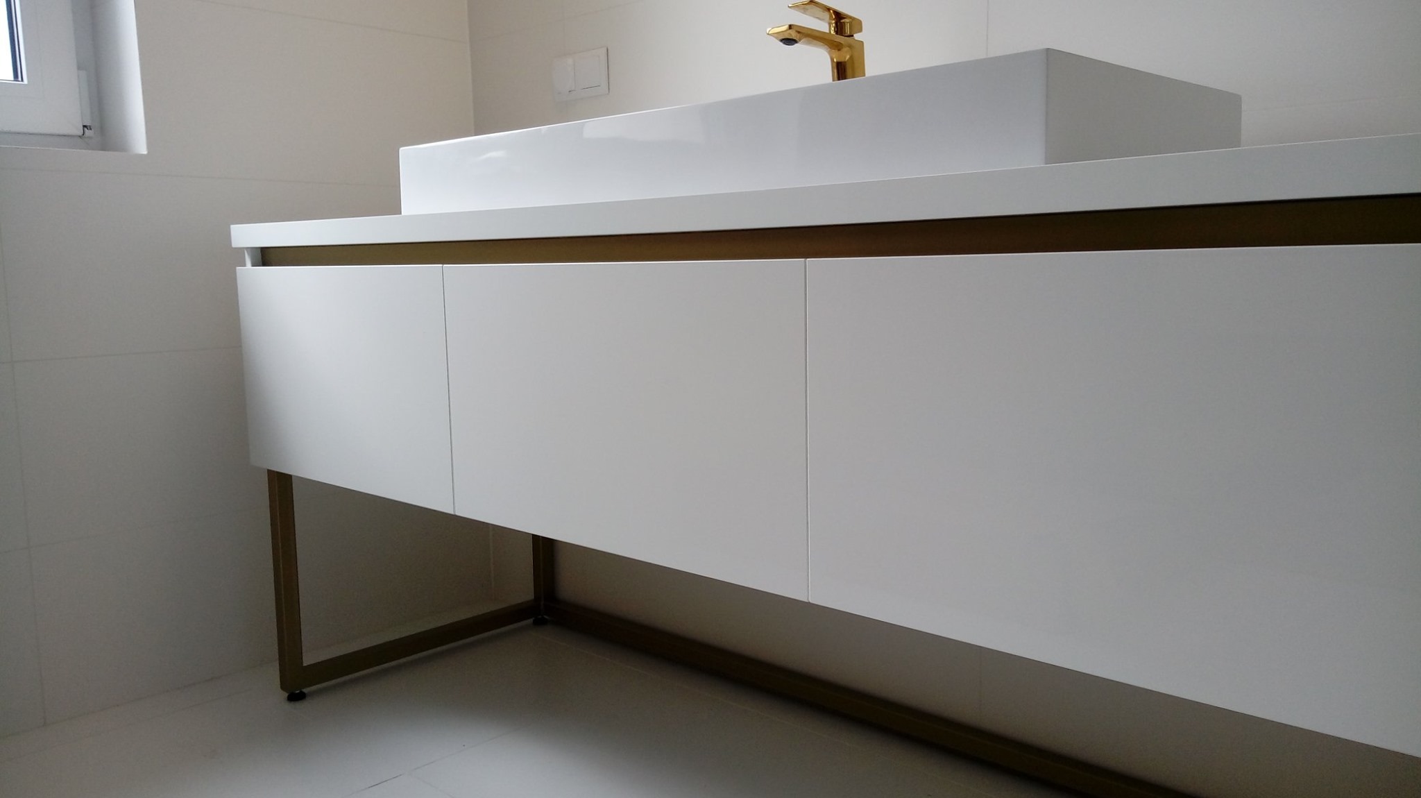 Nowoczesna biała szafka łazienkowa z trzema frontami i złotymi nogami, na której stoi prostokątna umywalka z baterią w kolorze złotym, na tle białych płytek.