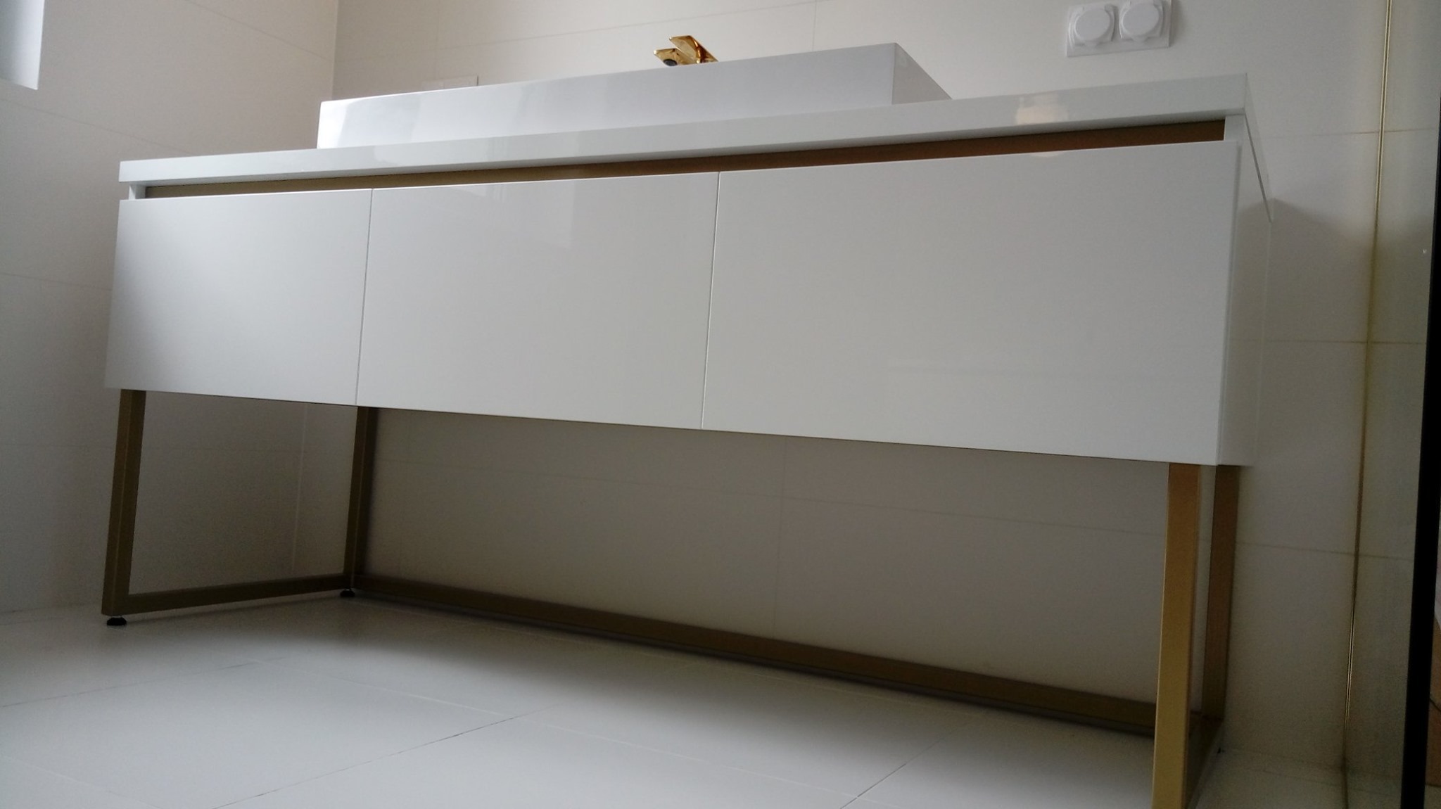 Biała szafka łazienkowa z trzema frontami i złotymi nogami, stojąca na białych płytkach, widok z dołu