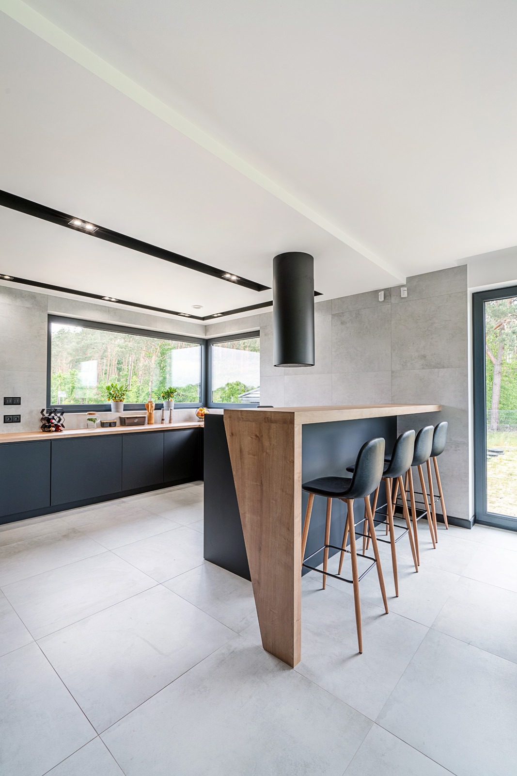 Nowoczesna kuchnia z wyspą i czterema hokerami, czarne szafki dolne z drewnianym blatem, szare płytki ścienne i podłogowe, okno z widokiem na zieleń.