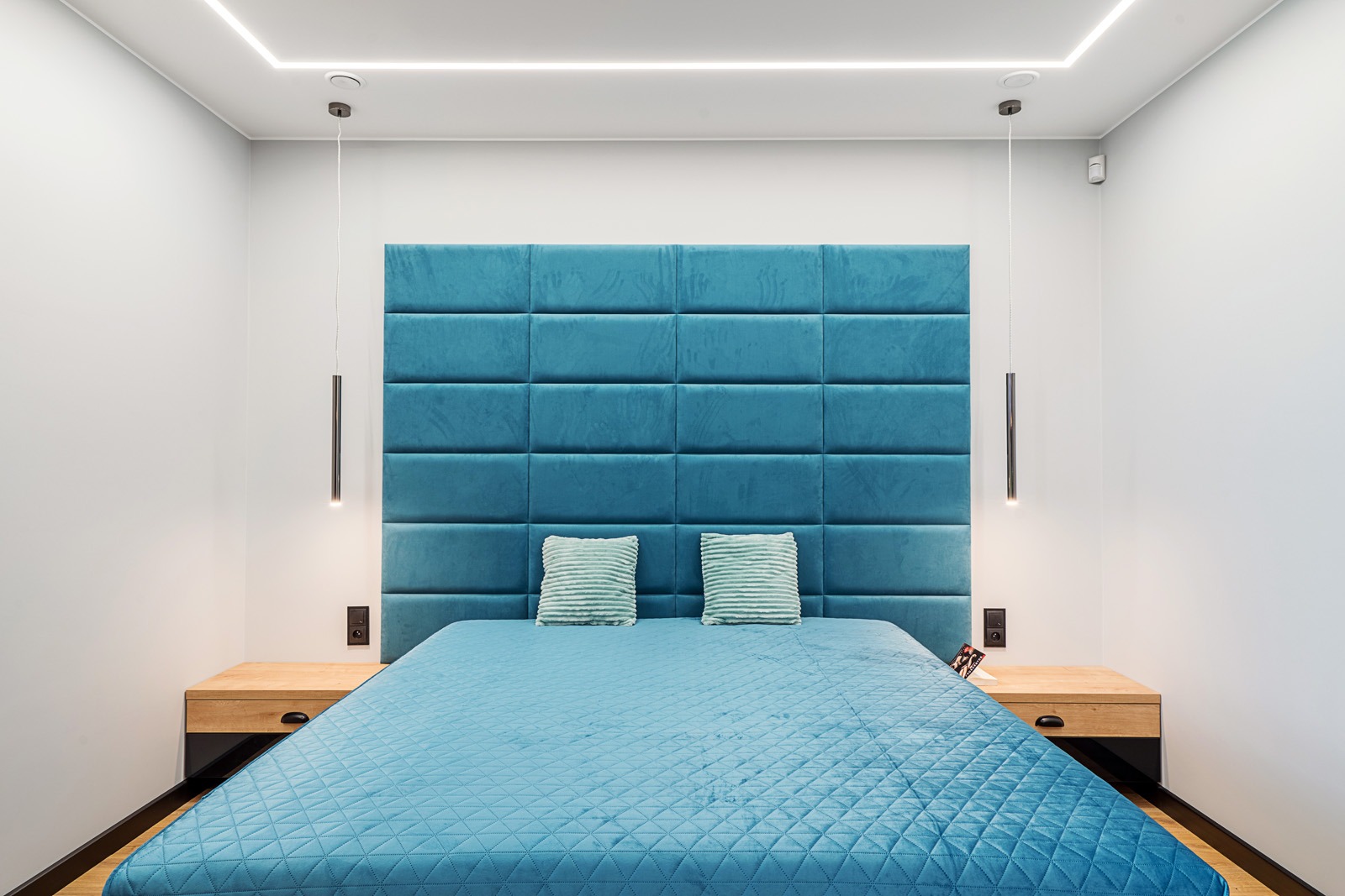 Nowoczesna sypialnia z turkusowym panelem tapicerowanym jako wezgłowie łóżka, minimalistyczne lampy wiszące i drewniane szafki nocne.