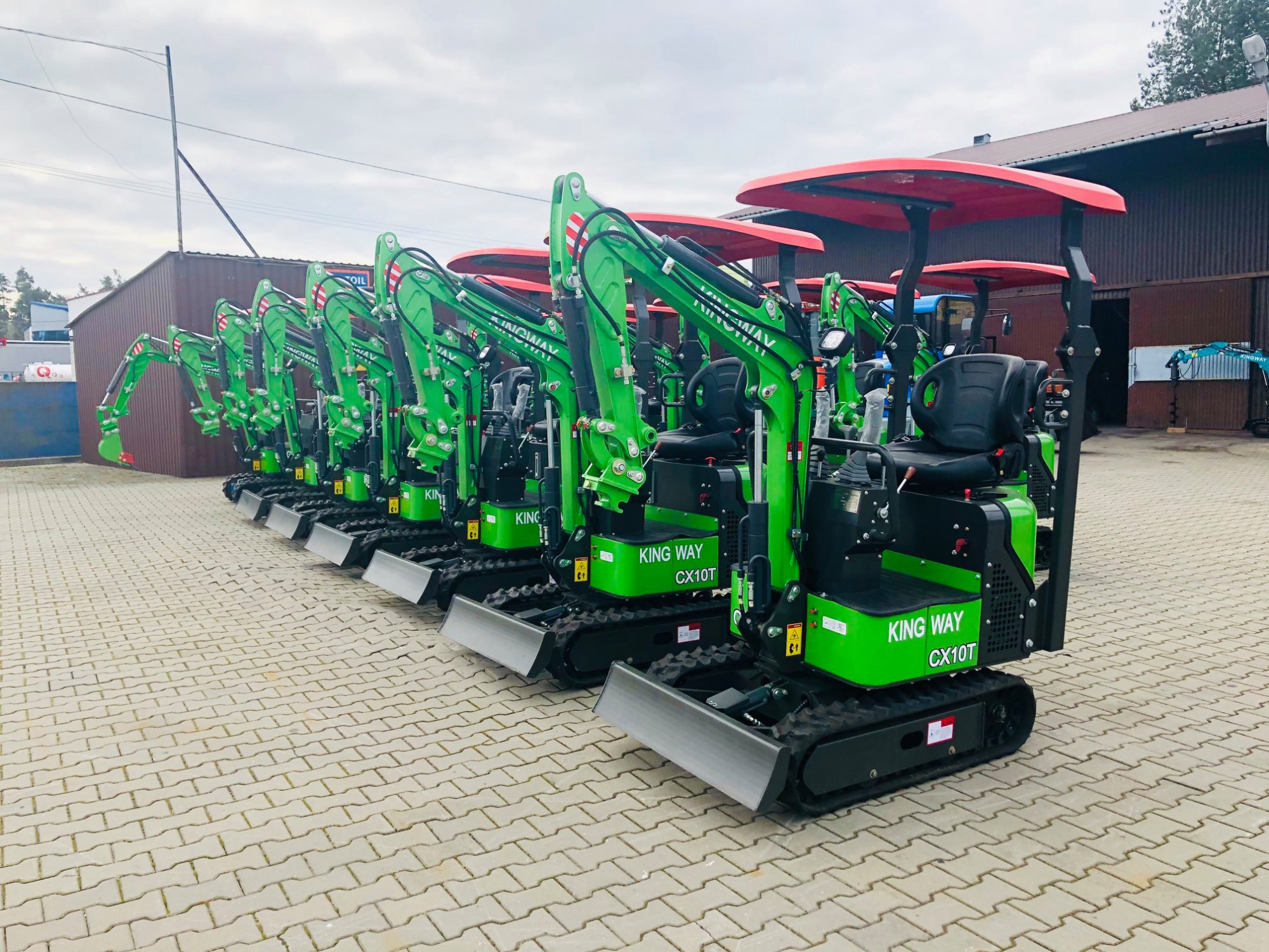Rząd nowych, zielonych mini-ładowarek teleskopowych King Way CX10T z czerwonymi daszkami, ustawionych na brukowanej powierzchni przed budynkiem.