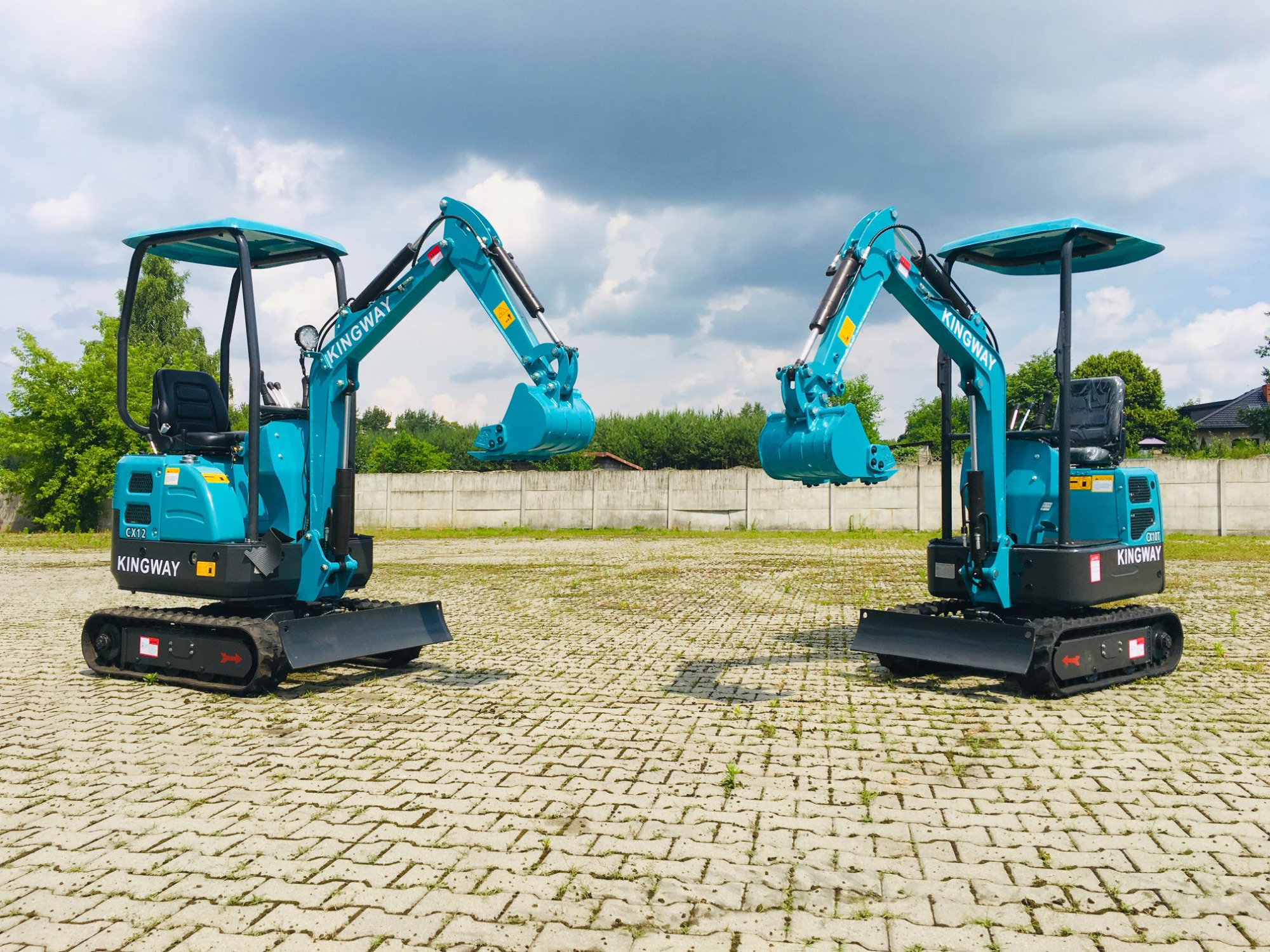 Dwa identyczne, turkusowe mini-koparki marki Kingway, ustawione obok siebie na placu wyłożonym kostką brukową, z widocznymi łyżkami skierowanymi ku sobie i tłem z zieleni oraz betonowego muru.