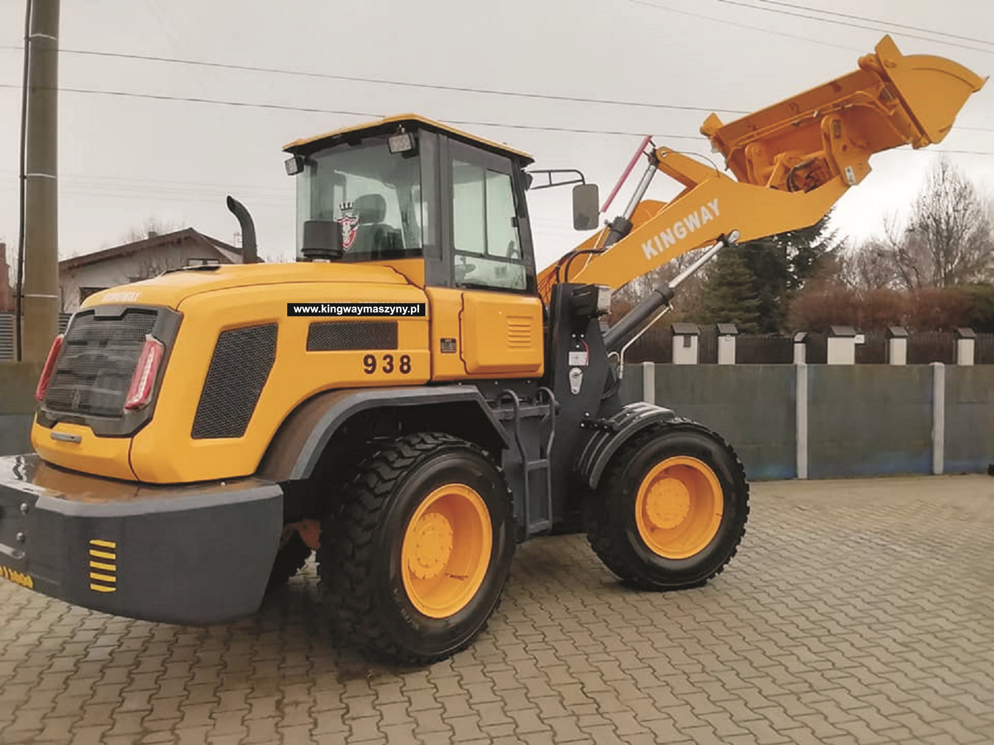 Żółta maszyna budowlana Kingway 938 z podniesioną łyżką, stojąca na kostce brukowej przed szarym ogrodzeniem.