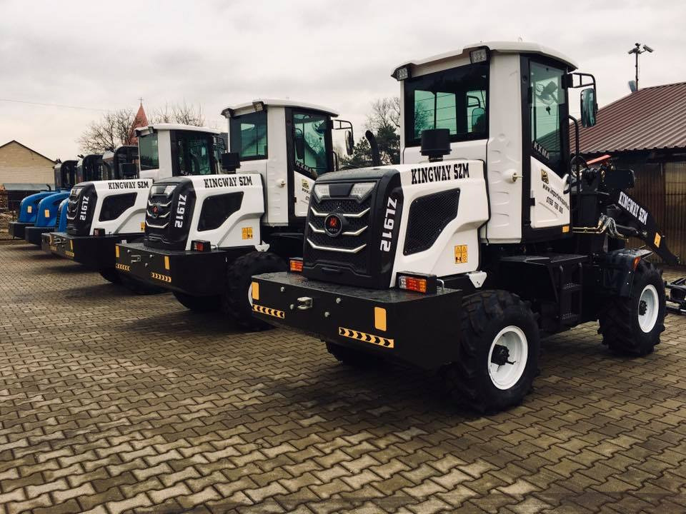 Szereg białych maszyn budowlanych KINGWAY SZM ZL912 ustawionych na brukowanej powierzchni, widoczne budynki w tle i pochmurne niebo.