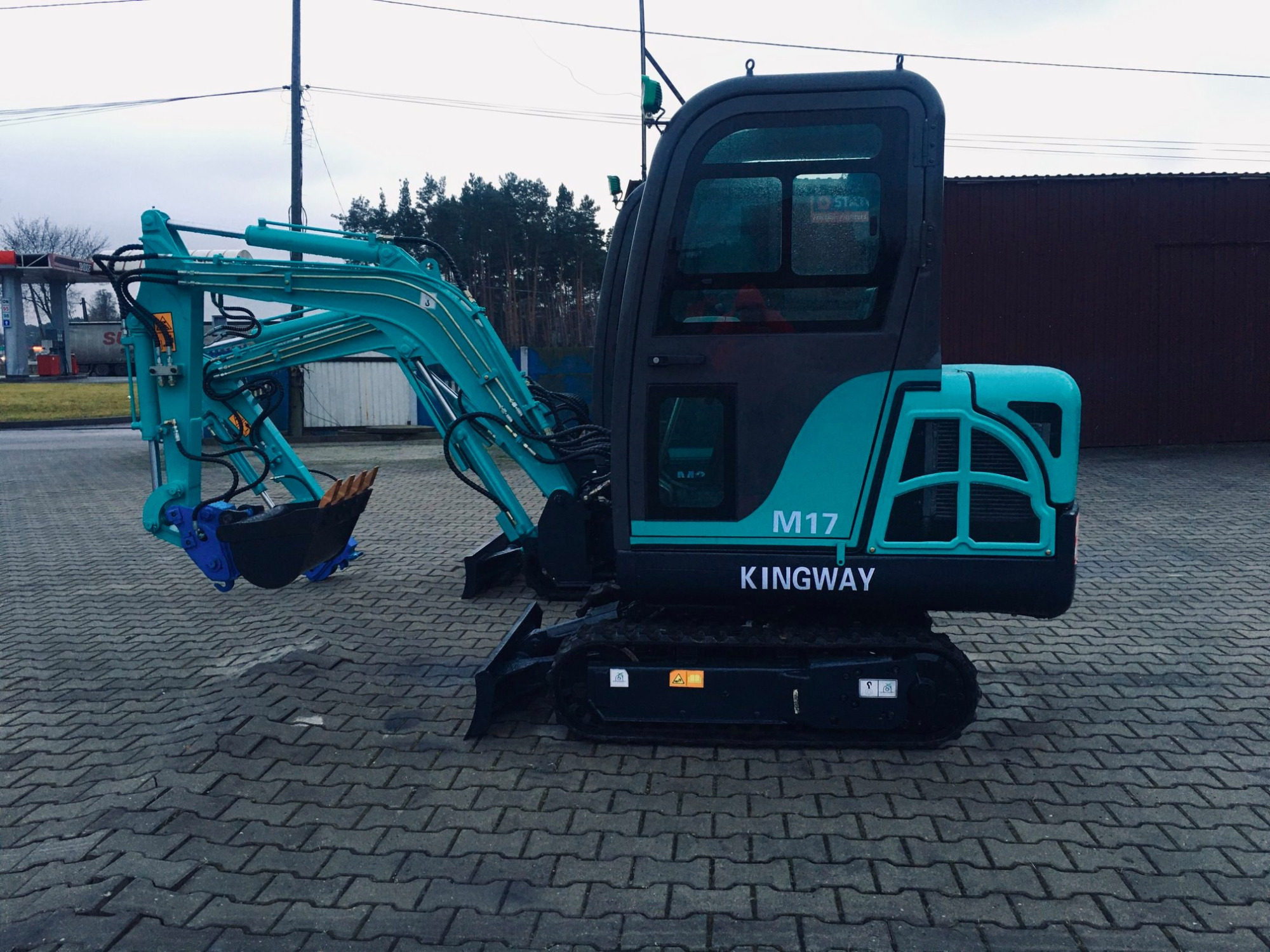 Niebiesko-czarna mini koparka Kingway M17 na brukowanym placu, z widoczną stacją benzynową w tle.
