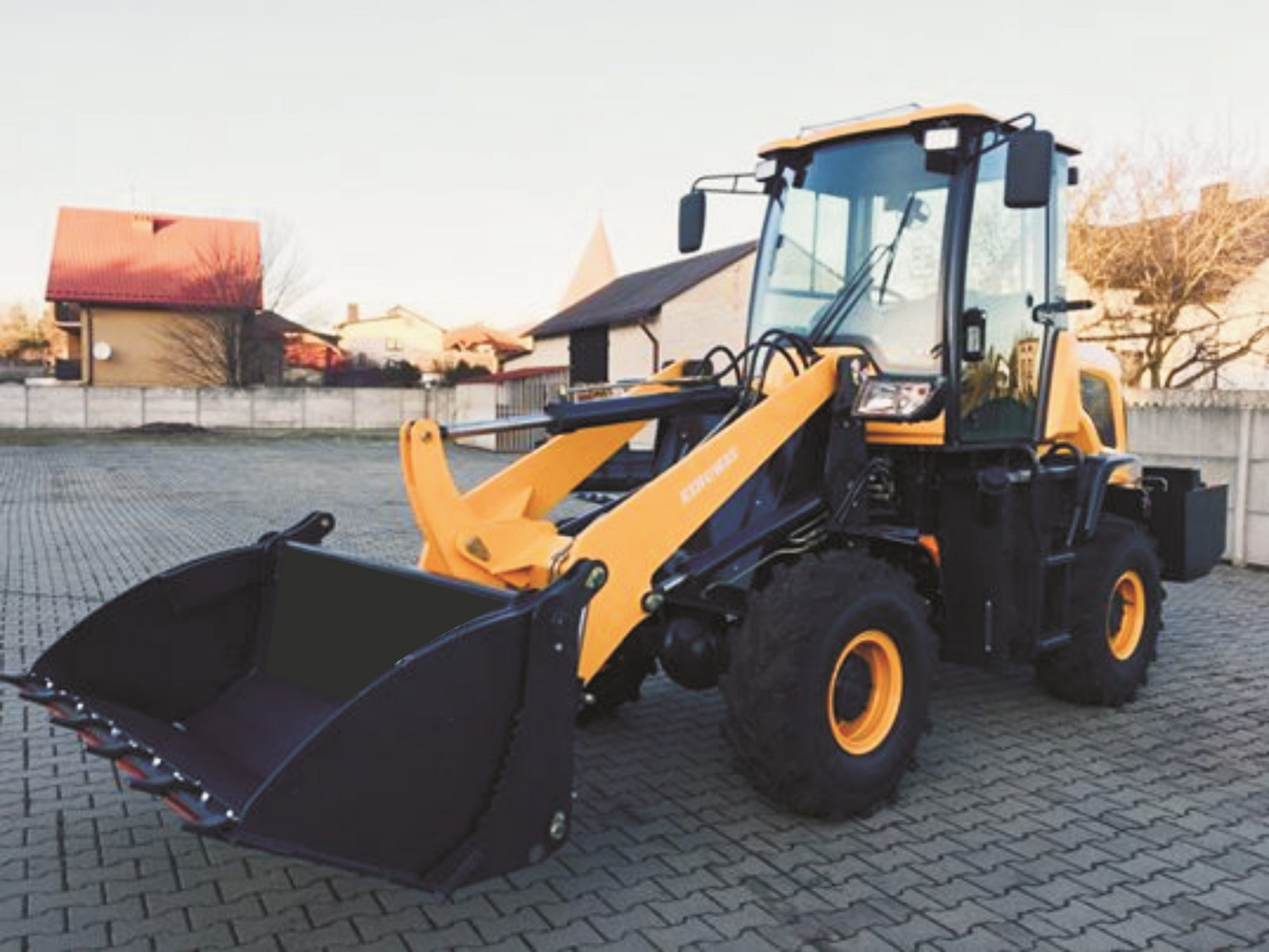 Żółto-czarna maszyna budowlana z łyżką na kostce brukowej, w tle zabudowa jednorodzinna i ogrodzenie.