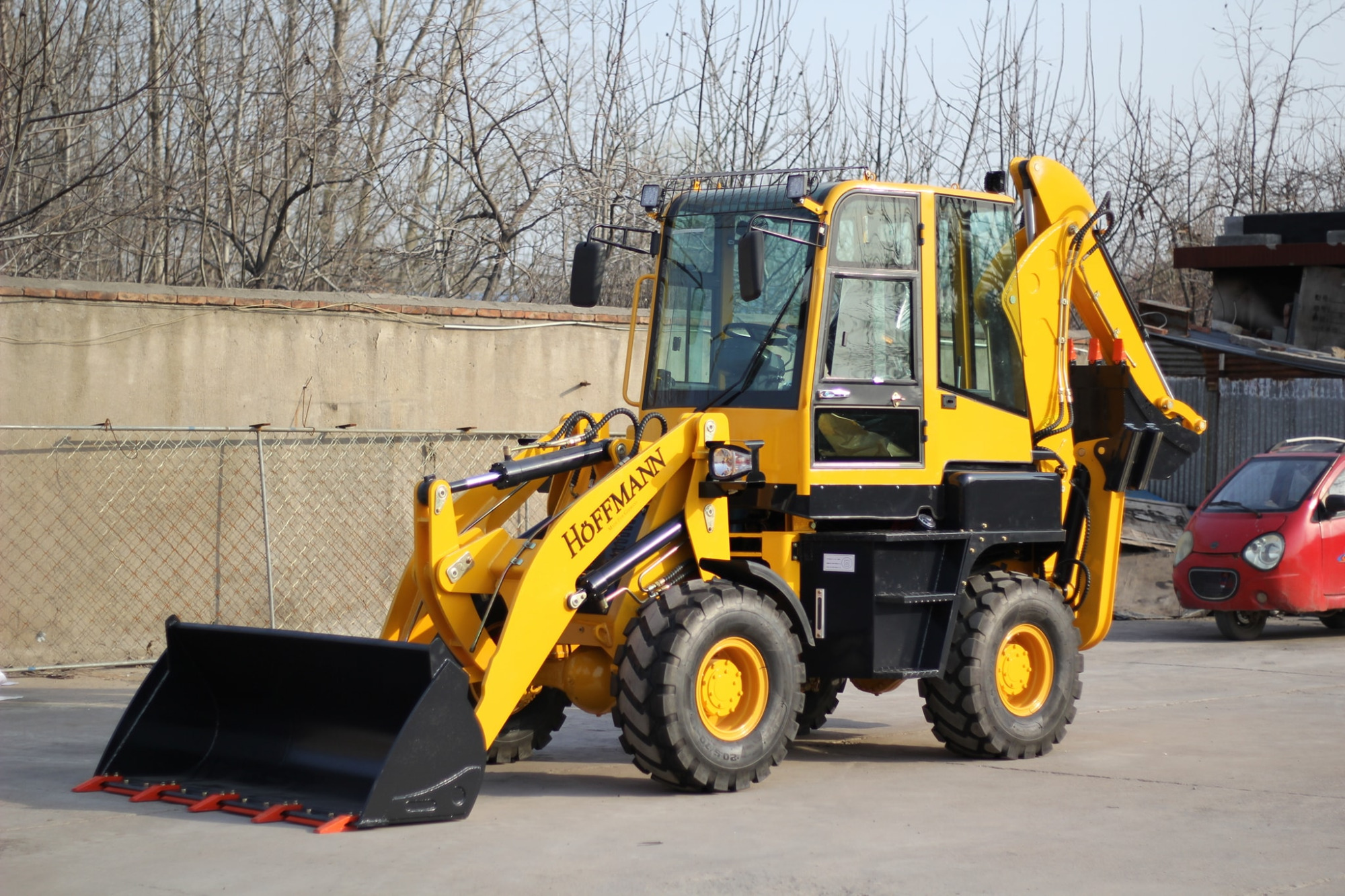 Żółta maszyna budowlana marki Hoffmann z czarną łyżką i pomarańczowymi zębami, zaparkowana na betonowym placu, z drzewami bez liści i czerwonym samochodem w tle.