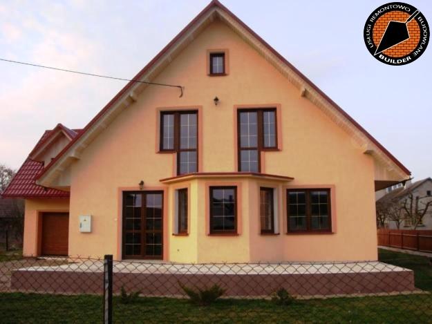 Żółty dom jednorodzinny z ciemnobrązowymi oknami i czerwonym dachem, widoczny zza siatki ogrodzeniowej, z logo firmy budowlanej w prawym górnym rogu.