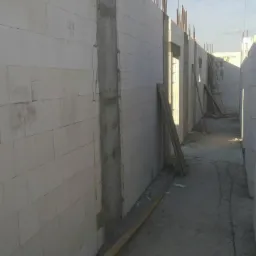 Wznoszenie ścian budynku z bloczków betonowych z widocznymi zbrojeniami pionowymi i częściowo wylane słupy betonowe.