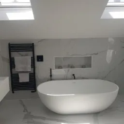 Nowoczesna łazienka z wolnostojącą wanną, płytkami imitującymi marmur, czarną armaturą i grzejnikiem drabinkowym, oświetlona światłem z okien dachowych.