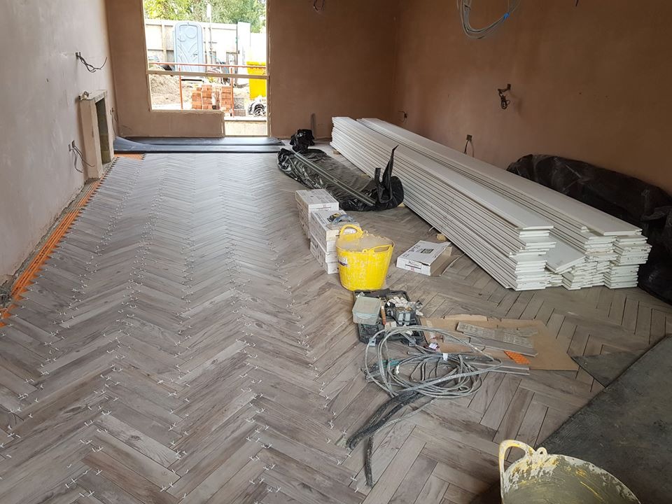 Układanie parkietu w jodełkę w pomieszczeniu w stanie surowym, widoczne białe listwy przypodłogowe, żółte wiadro i narzędzia pracy.