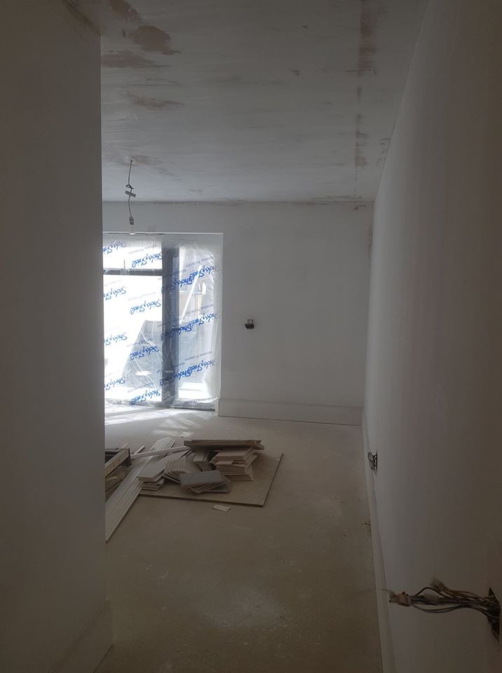 Przygotowane do malowania wnętrze z zabezpieczonym oknem folią, widoczne listwy przypodłogowe i wystające przewody elektryczne.
