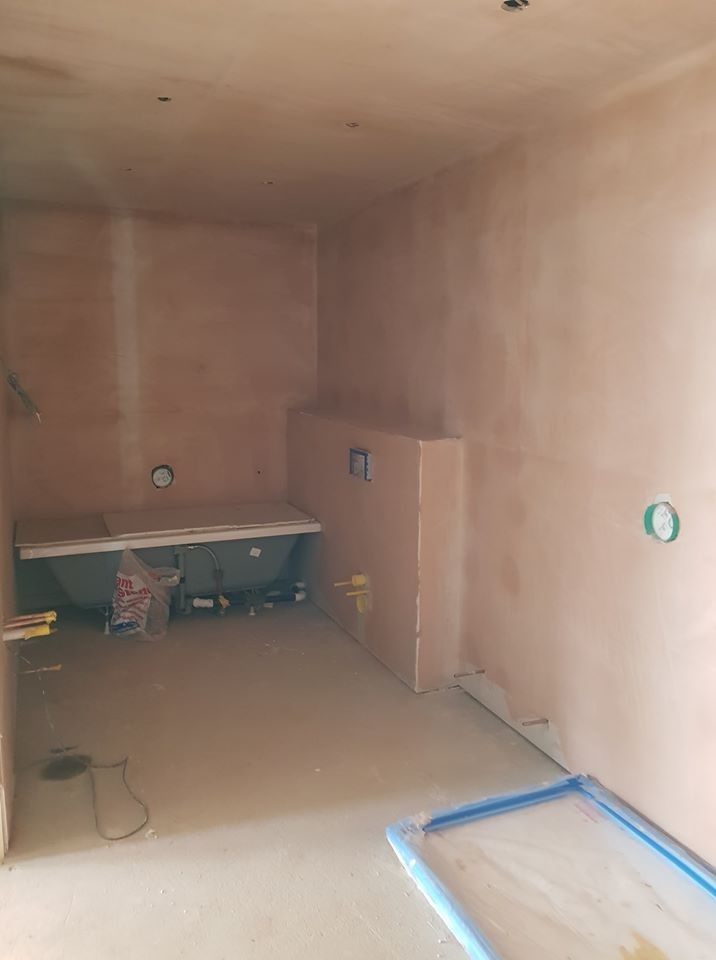 Surowa łazienka w trakcie remontu, ściany pokryte świeżym tynkiem, widoczna wanna z podłączoną instalacją, podłoga częściowo zabezpieczona folią malarską, puszki elektryczne na ścianach.