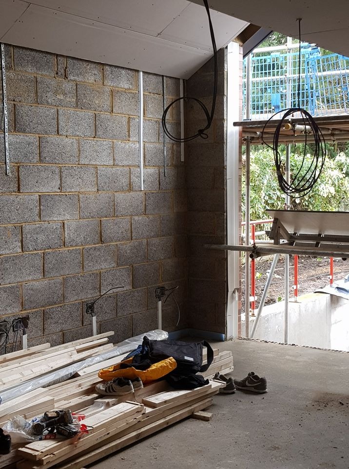 Wnętrze w trakcie remontu: surowa ściana z betonowych bloczków, wystające kable elektryczne, przygotowane deski i narzędzia na podłodze, widok na rusztowanie i zieleń na zewnątrz.