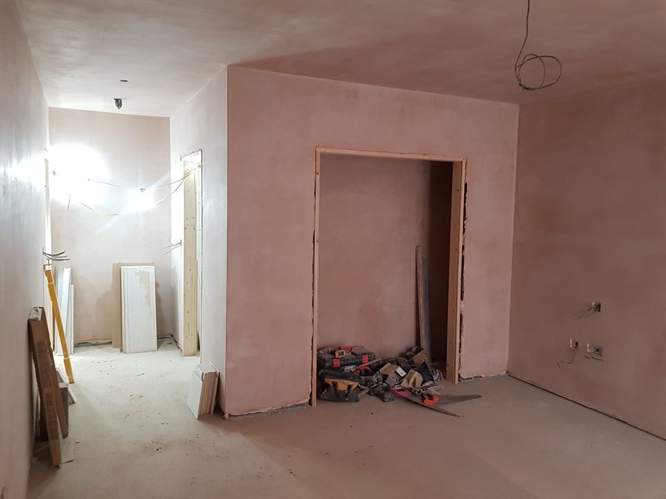 Wnętrze w trakcie prac wykończeniowych: świeżo otynkowane ściany w odcieniach beżu, widoczne narzędzia i kable, przygotowane ościeżnice drzwiowe.
