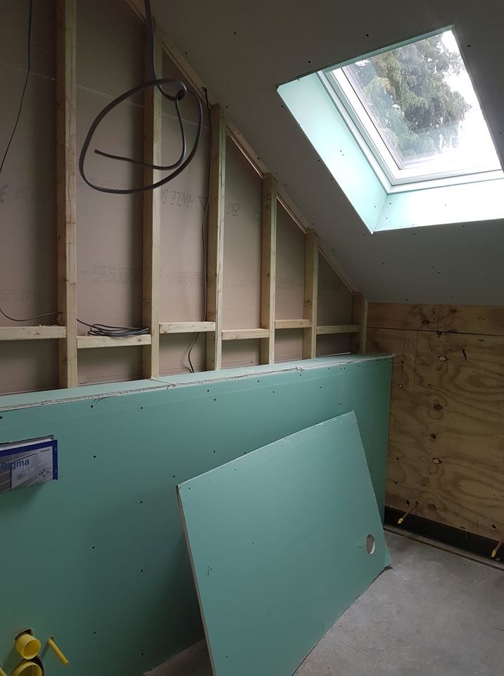 Poddasze w trakcie remontu: widoczne płyty gipsowo-kartonowe, drewniana konstrukcja szkieletowa, okno dachowe i instalacje elektryczne, z żółtymi elementami instalacji wodnej w dolnym rogu.