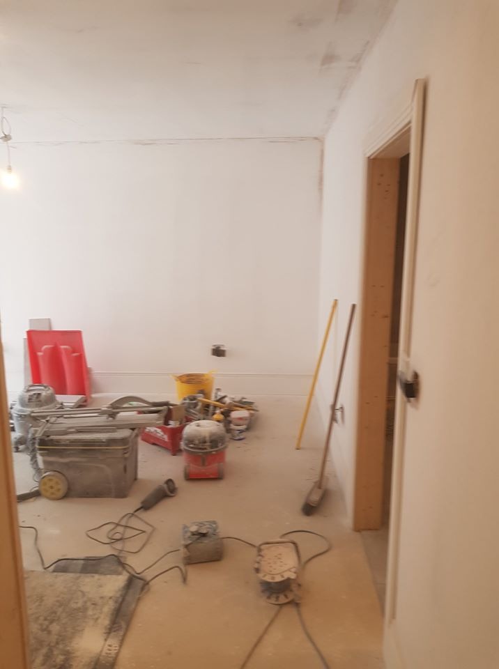 Pokój w trakcie remontu: widoczne narzędzia glazurnicze, szlifierka do gładzi, odkurzacz przemysłowy, żółte wiadro i czerwone płyty, ściany przygotowane do malowania, sufit z widocznymi śladami...
