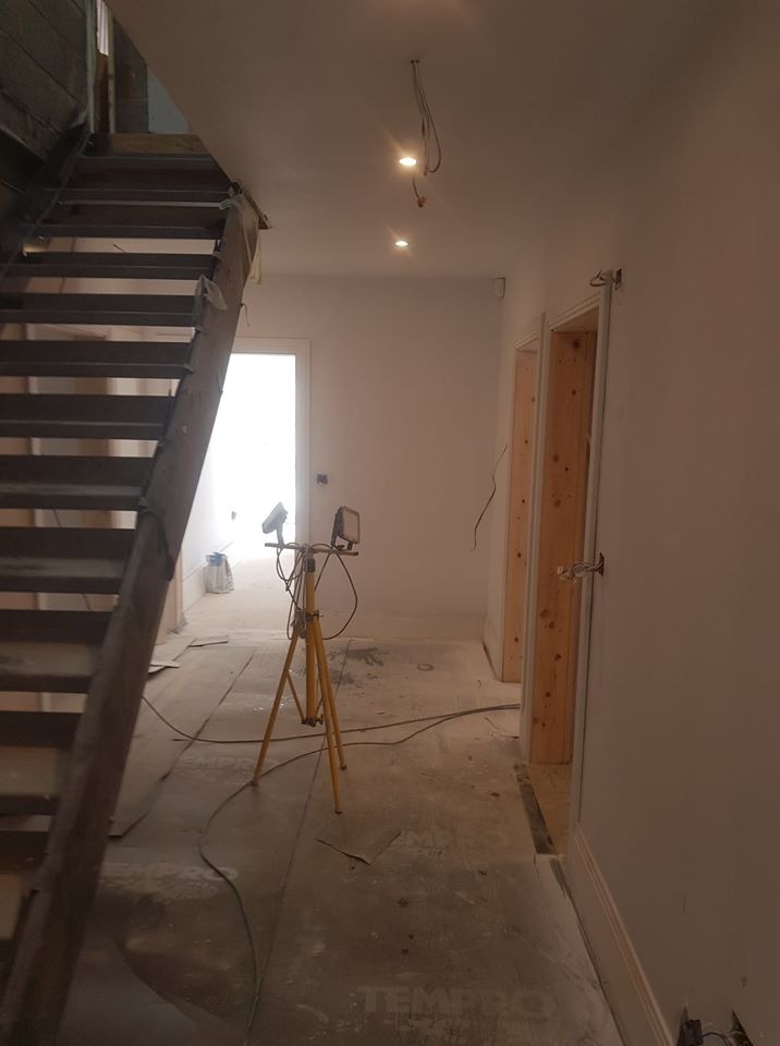 Korytarz w trakcie remontu, widoczne schody drewniane, przygotowane ościeżnice drzwiowe, podłoga zabezpieczona folią, lampa budowlana na trójnogu, surowe ściany przygotowane do malowania, widoczne...